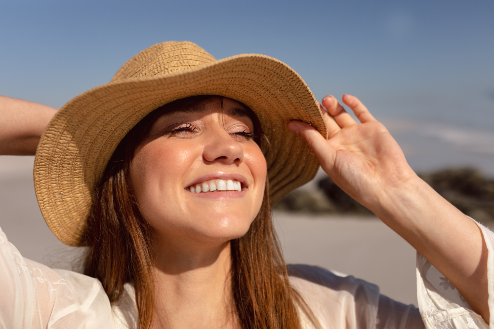 muher branca, de cabelo comprido, usando um chapéu na praia e sorrindo
