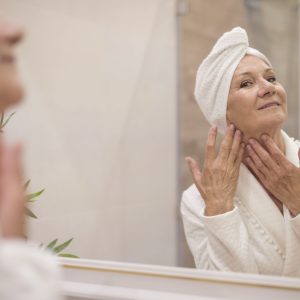 mulher mais velha em um pós banho tocando a pele e se olhando no espelho