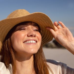 muher branca, de cabelo comprido, usando um chapéu na praia e sorrindo