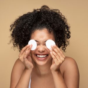 mulher negra, com cabelo preso segurando dois algodões nos olhos e sorrindo