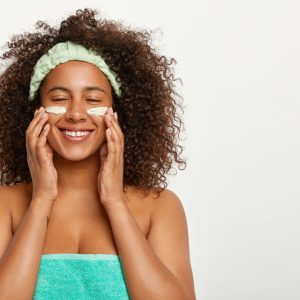 mulher negra, usando uma toalha verde, usando um produto de beleza na pele do rosto